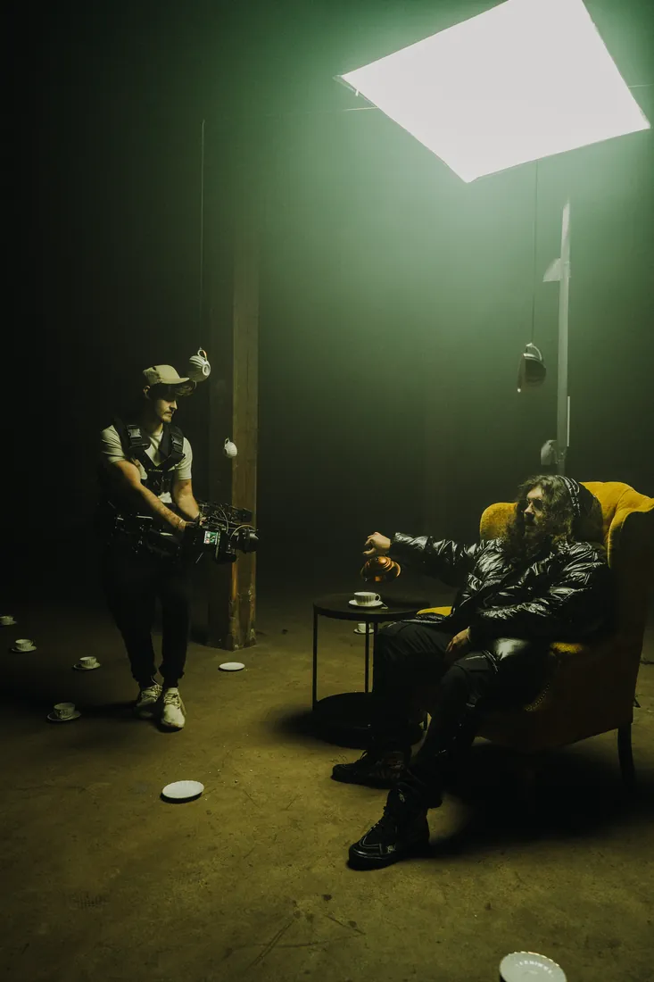 A Steadicam operator circles an artist seated in an armchair in a hazy, dimly lit room.